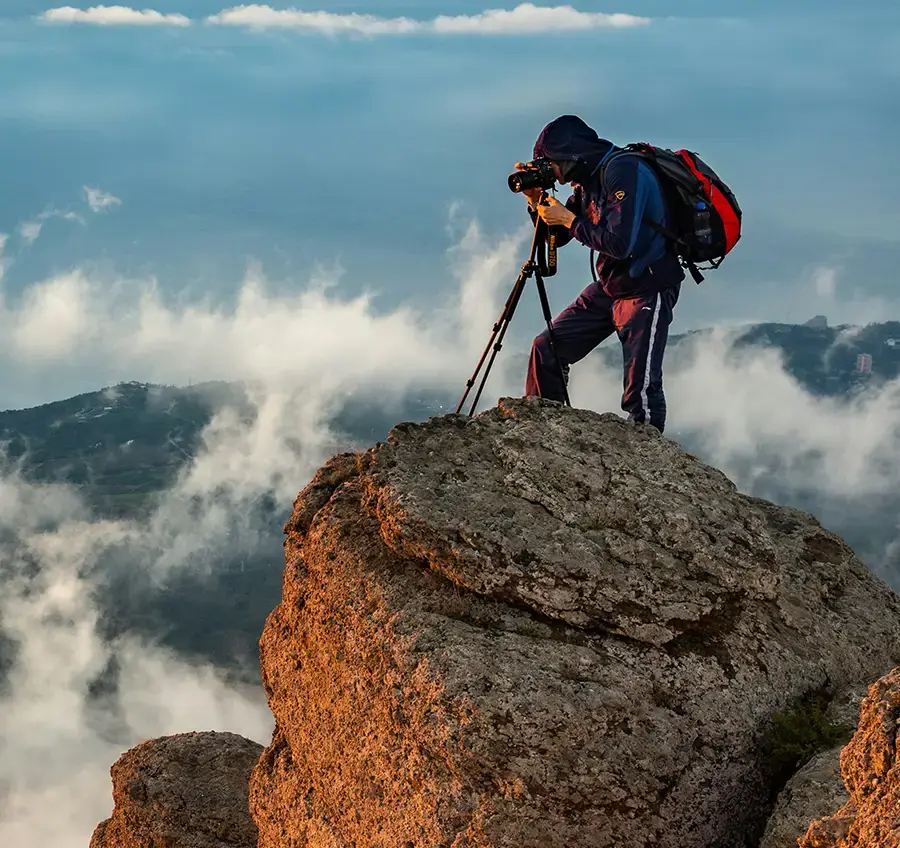 Ein Fotograf steht mit Stativ und Kamera auf einem großen Stein in den Bergen